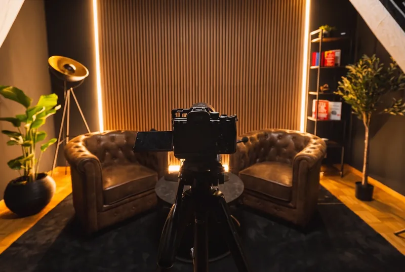 Videopodcast Studio in Bern-Liebefeld: Kamera mit ausgeklapptem Monitor vor den zwei Sesseln, dahinter die Holzlamellenwand mit Lichtkanten