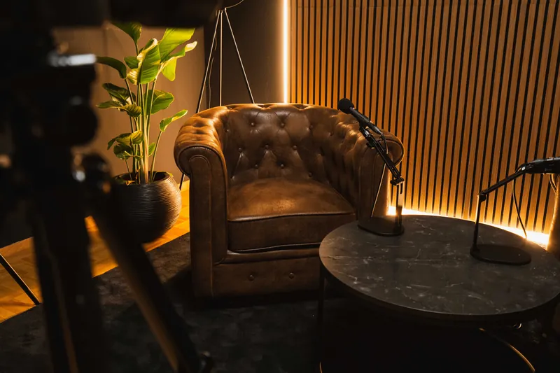 Chesterfield-Sessel mit Mikrofon am runden Tisch, daneben die Holzlamellenwand mit Lichtkante