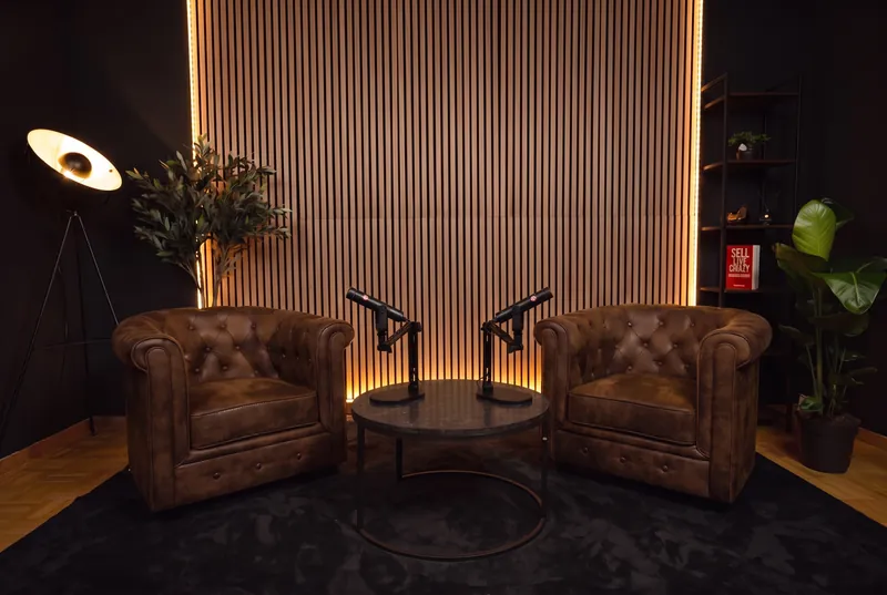 Podcast Studio in Bern-Liebefeld: zwei Chesterfield-Sessel und ein runder Tisch mit zwei Mikrofonen vor der beleuchteten Holzlamellenwand